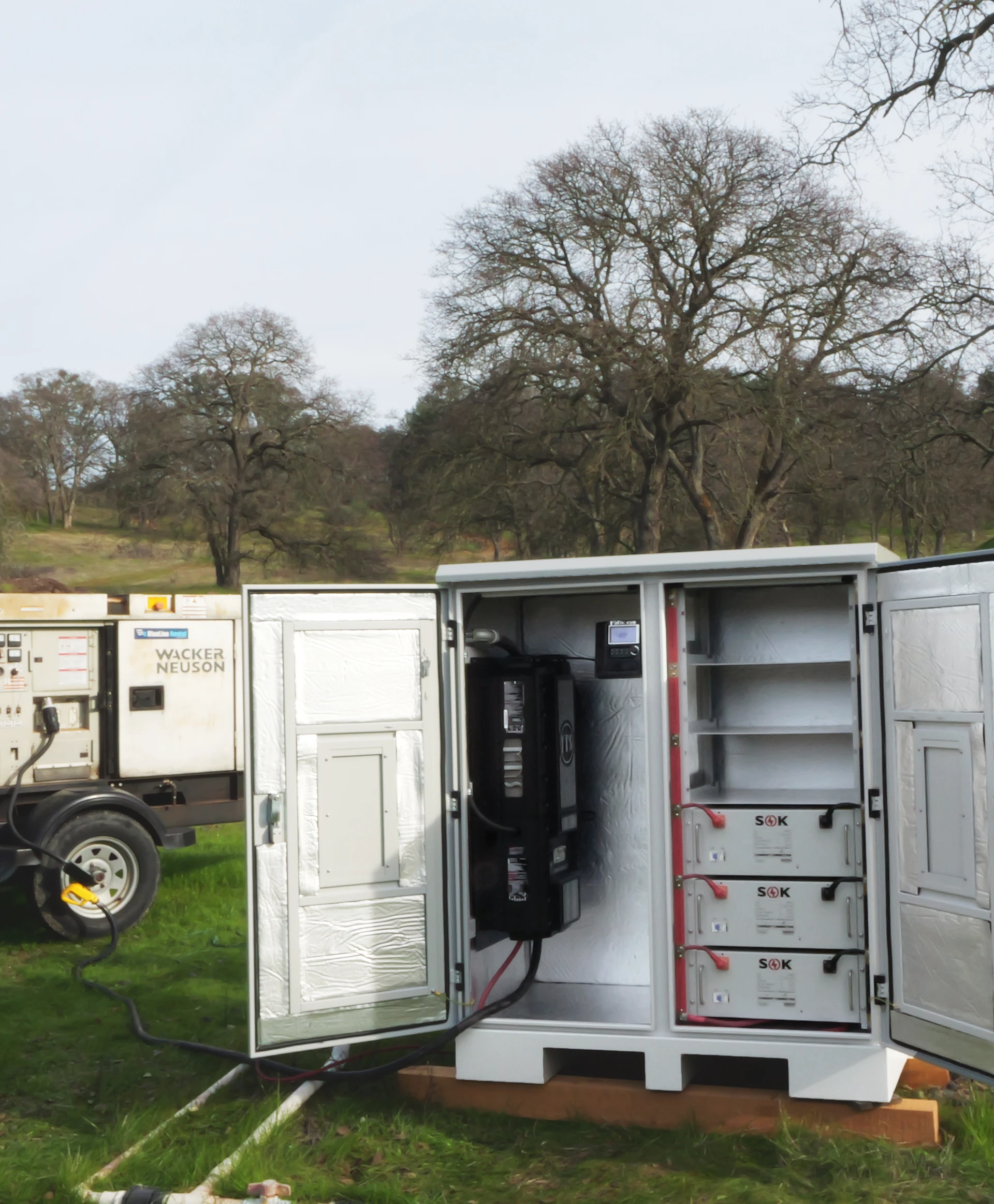 Photo of an inverter and batteries in a cabinet. The cabinet is on a pallet in the middle of a field with trees all around. 